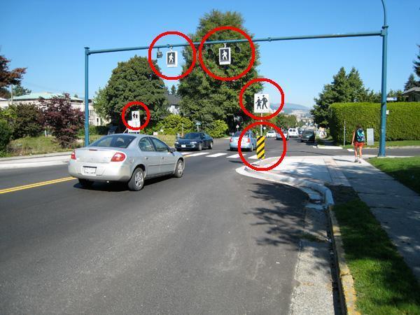 Image of a crossing in Vancouver features kerb extensions and zebra stripes, signage and amber pedestrian activated lights, hung overhead, to help motorists identify this as a crossing. Also, there are hazard signs to warn motorists that the road narrows between the kerb extensions 
