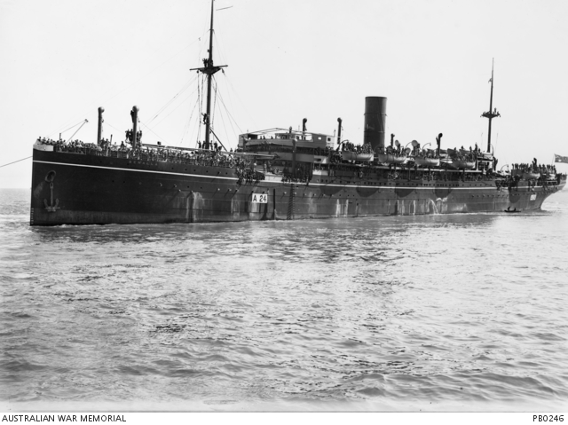 A large, dark coloured ship with a single funnel crowded with troops, departing from the wharf. A small rowing boat is near the stern of the ship.